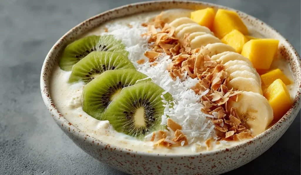 Coconut smoothie bowl with kiwi slices, mango, banana, and toasted coconut flakes in a white bowl on a gray countertop.