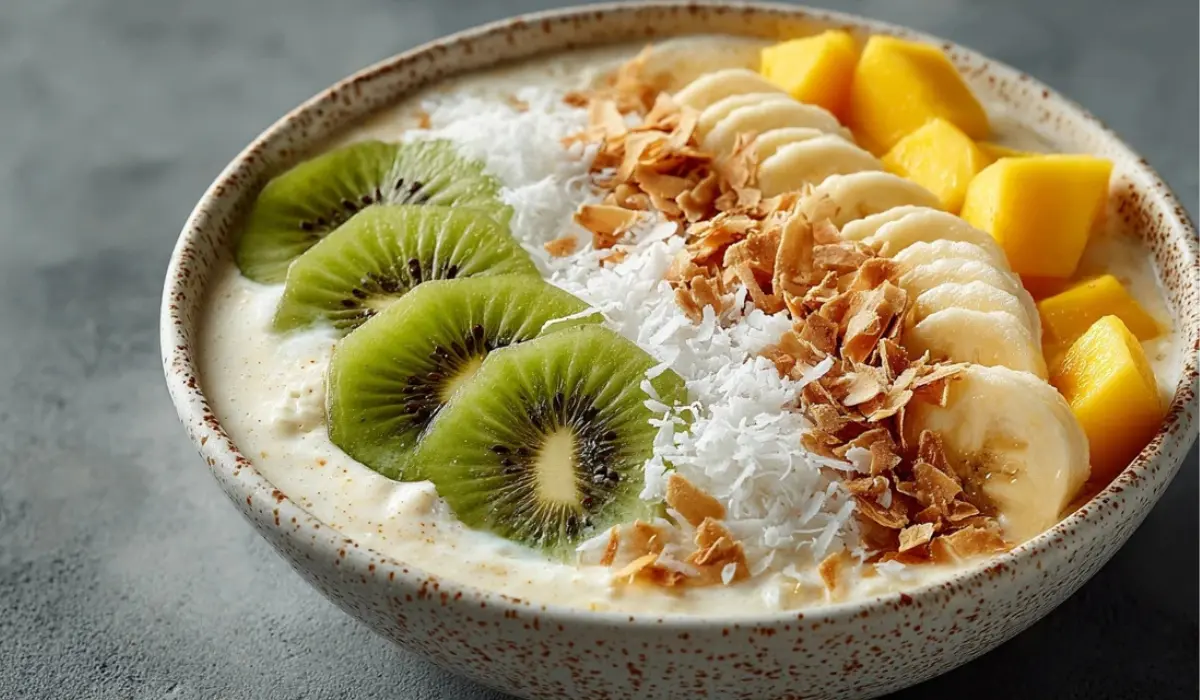 Creamy coconut smoothie bowl topped with mango, kiwi, banana slices, and toasted coconut flakes on a gray countertop.