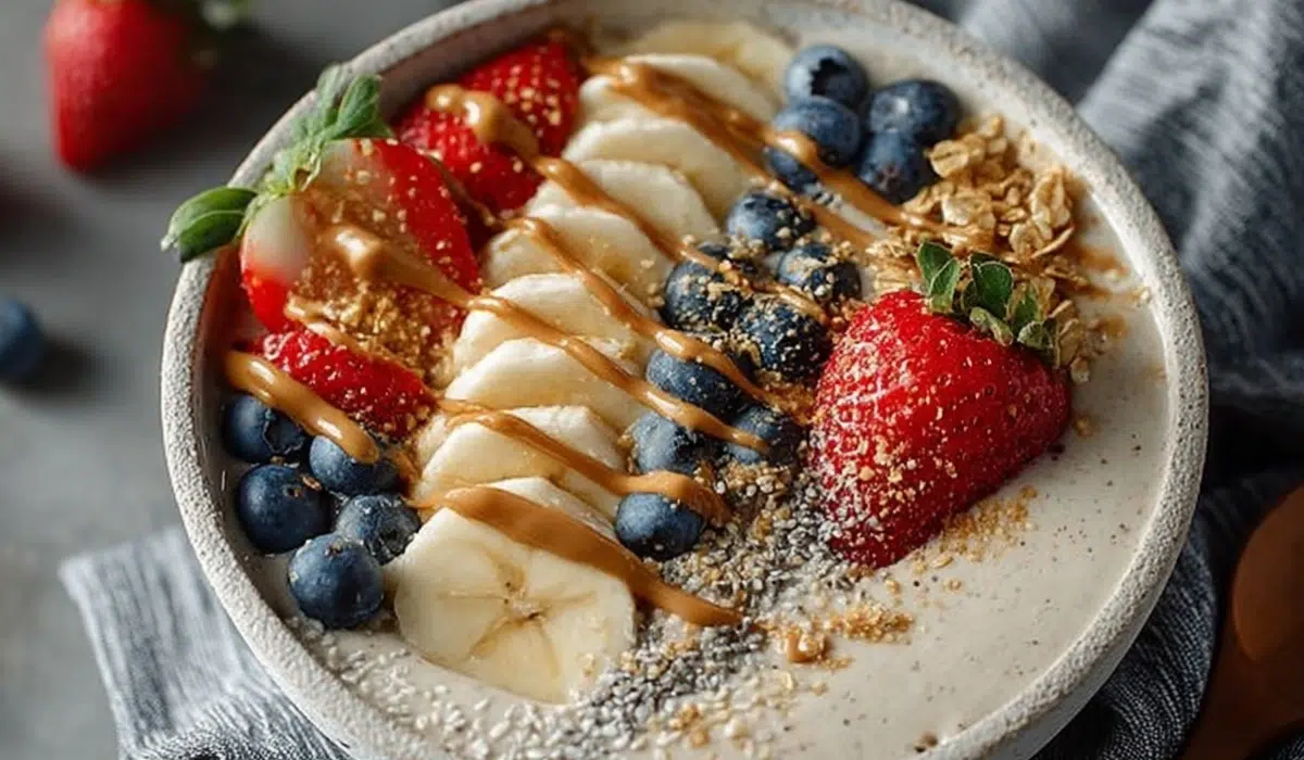 Overhead shot of a thick, creamy smoothie bowl in a white bowl on a gray kitchen countertop, topped with fresh fruits, granola, and a drizzle of nut butter.