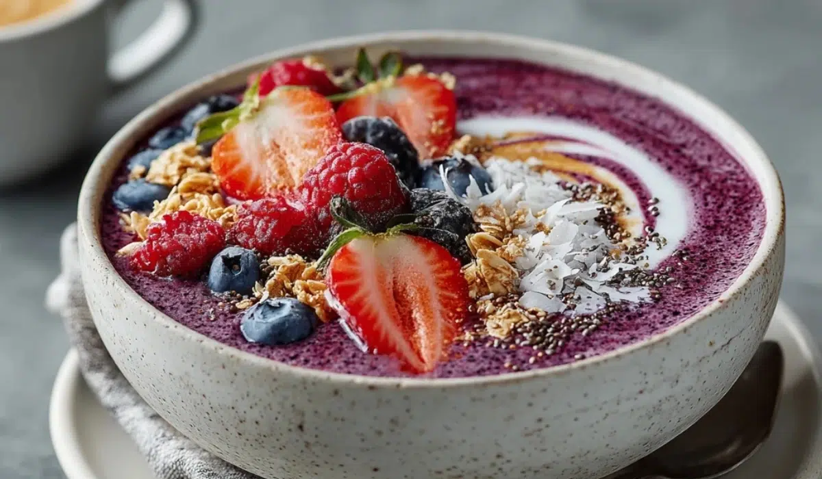 Thick mixed berry smoothie bowl in a white ceramic bowl on a gray countertop, topped with strawberries, blueberries, raspberries, granola, chia seeds, and coconut flakes.