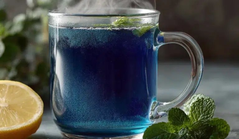Glass mug of deep blue natural Mounjaro tea with lemon wedge on the side, steam rising, fresh mint leaves scattered on a gray countertop.