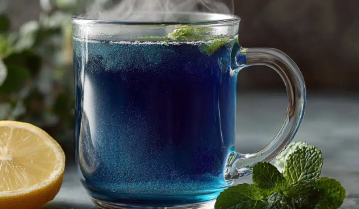 Glass mug of deep blue natural Mounjaro tea with lemon wedge on the side, steam rising, fresh mint leaves scattered on a gray countertop.