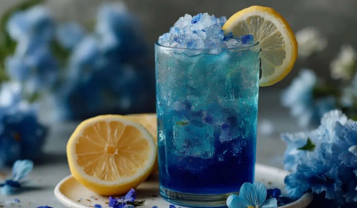 Glass of bright blue salt trick drink with lemon wedge on a white plate, gray kitchen countertop background.