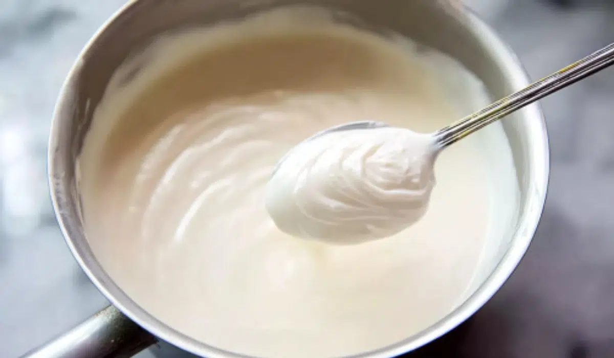 Overhead shot of creamy béchamel sauce in a white bowl on marble background, smooth and glossy texture, photographed in soft natural light.
