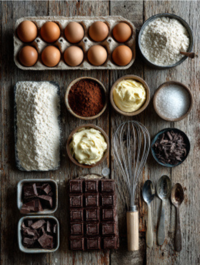 Organized flat lay of Chocolate Yule Log ingredients including eggs, cocoa powder, flour, mascarpone, cream, sugar, and chocolate chunks on a wooden surface.