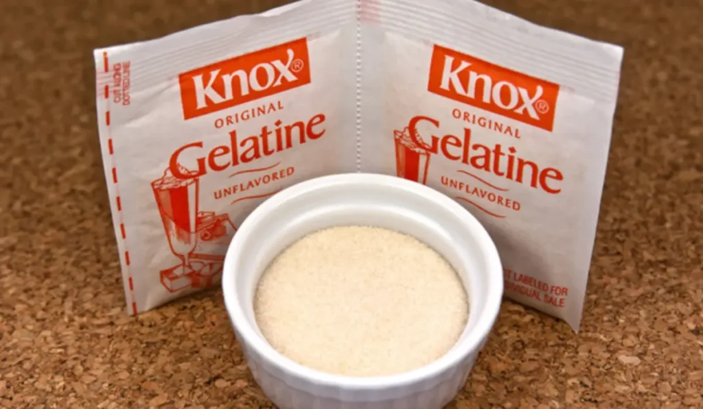 Knox unflavored gelatin packets placed on a marble kitchen counter beside a small bowl of clear gelatin powder and a glass of water.