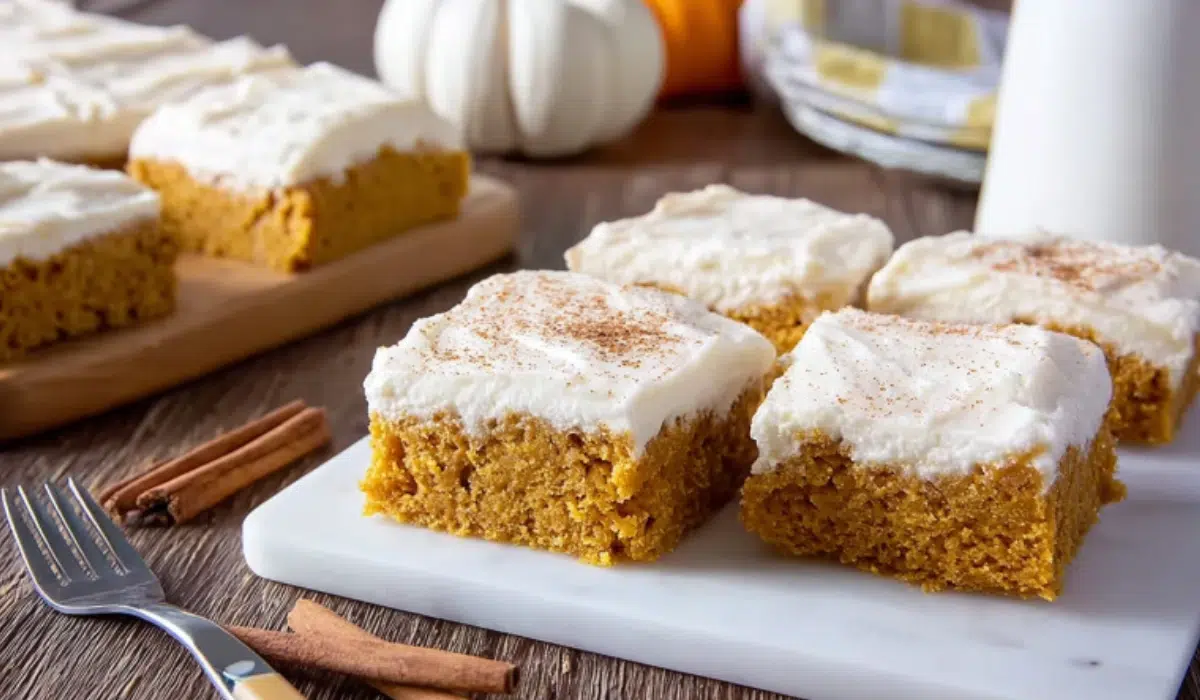 Moist pumpkin bars topped with smooth cream cheese frosting on a marble counter with cinnamon sticks and a fall cozy atmosphere.
