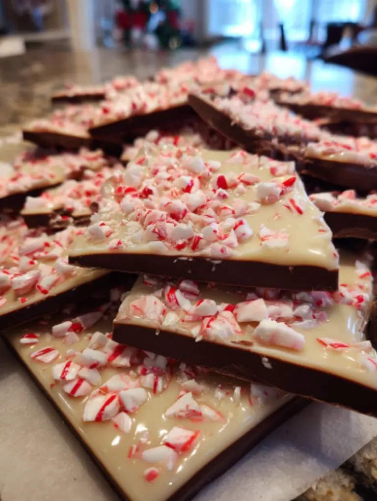Flat lay of peppermint bark pieces on a festive platter beside candy canes and red ribbons on a kitchen counter.