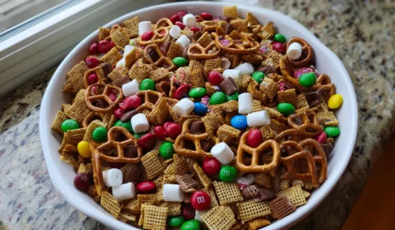Overhead photo of a festive Christmas Chex Mix in a white bowl on the kitchen counter, filled with pretzels, M&M’s, marshmallows, and sprinkles.