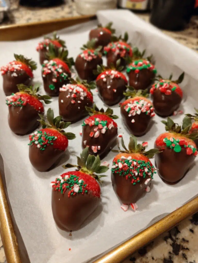 Homemade Christmas chocolate covered strawberries served on a holiday dessert board with cookies, peppermint bark, and truffles, displayed on a cozy kitchen counter.