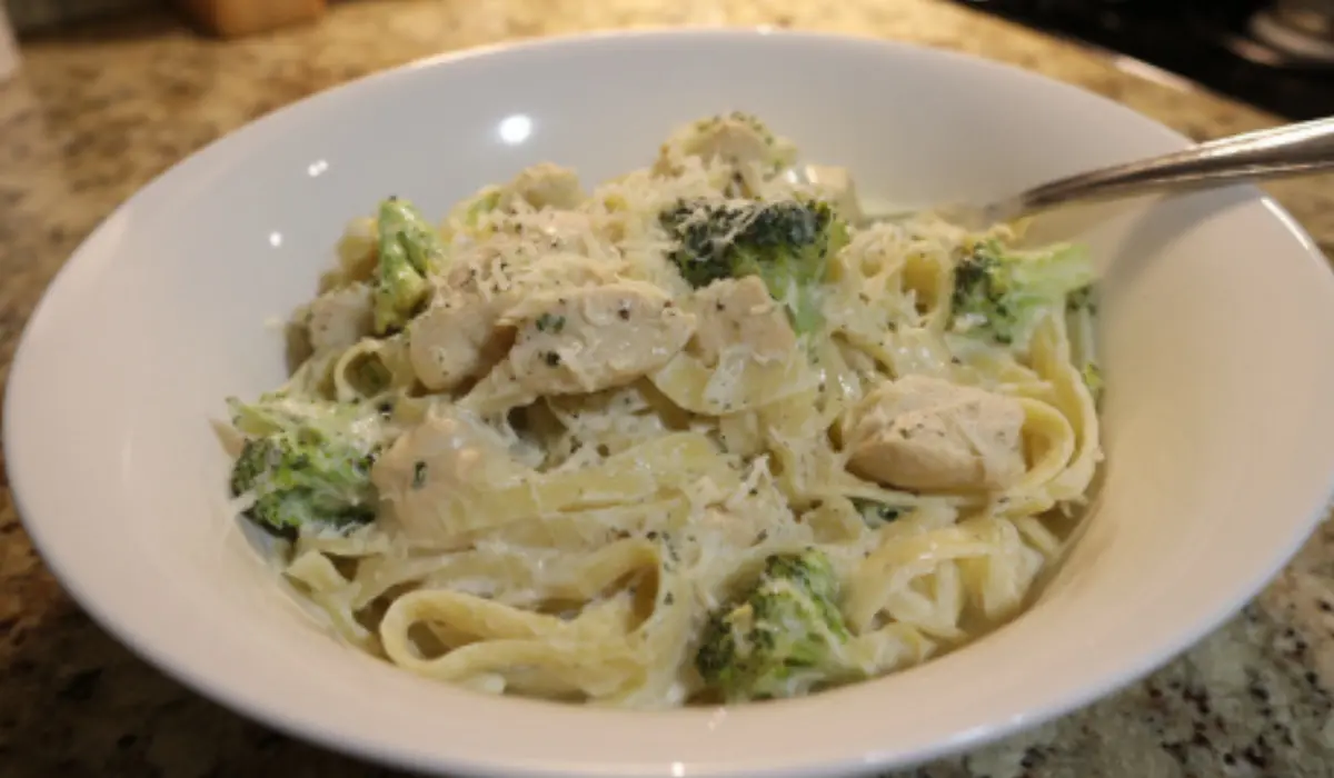 Flat lay of creamy chicken and broccoli fettuccine Alfredo served in a white bowl on a kitchen counter, topped with Parmesan and surrounded by simple home props.