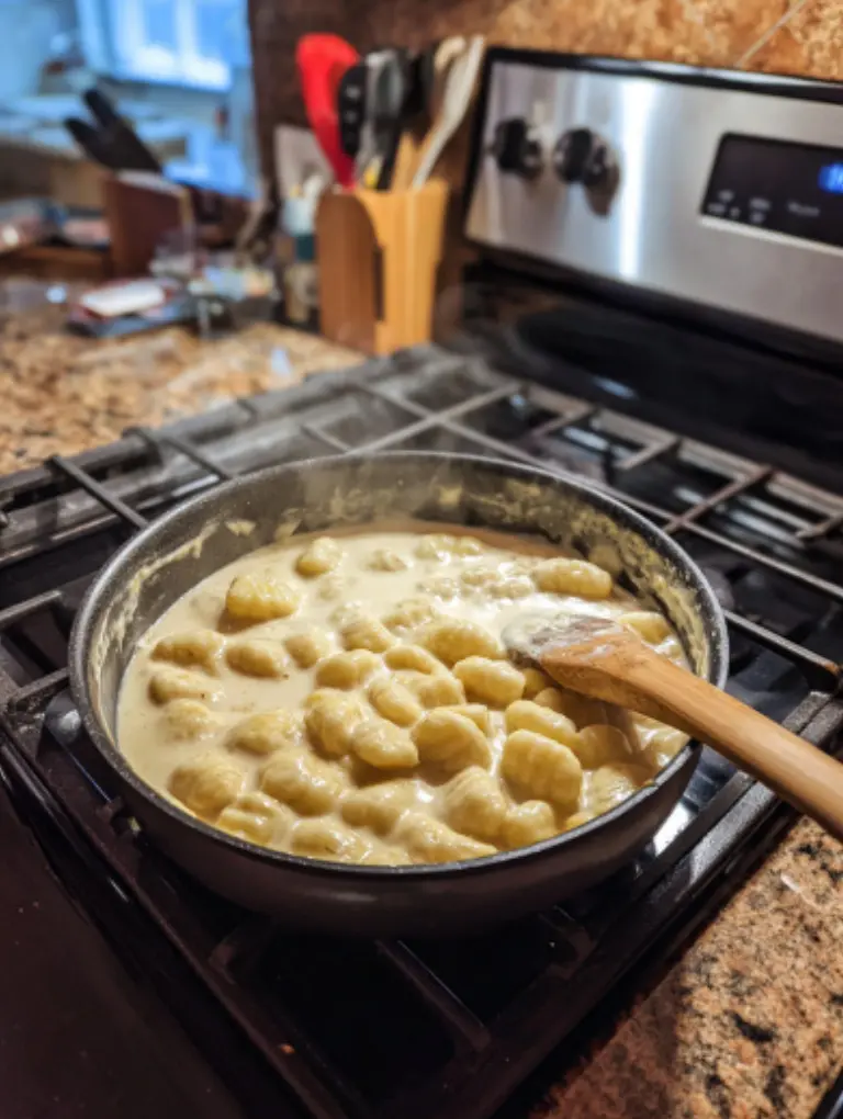 Flat lay of creamy gnocchi simmering in a skillet on the stove, sauce bubbling gently with a wooden spoon beside it.