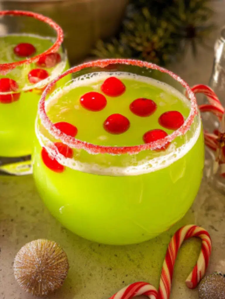 Glasses of green Grinch punch with red sugar rims and cherries on top, arranged on a kitchen counter with Christmas cookies and pine sprigs.