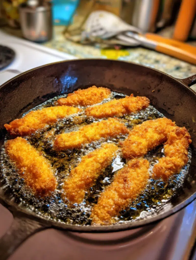 Golden honey-pepper chicken tenders frying in a cast-iron pan on the kitchen counter, oil bubbling around the crispy coating.