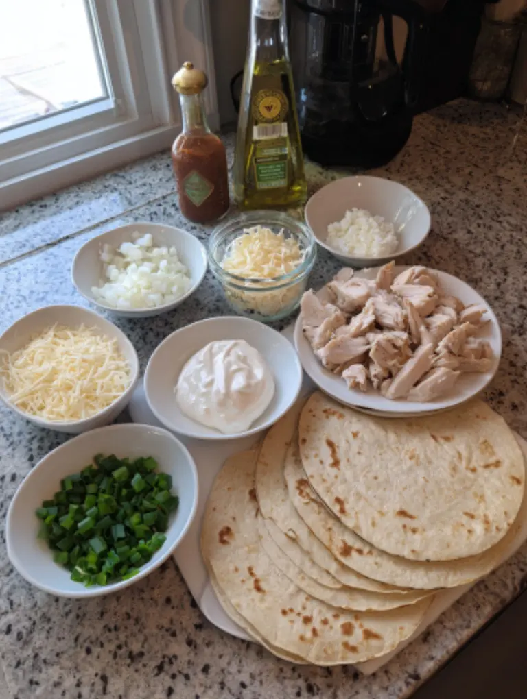 Flat lay of cooked chicken, minced garlic, mayonnaise, Greek yogurt, hot sauce, salt, tortillas, grated cheese, chopped green onion, and olive oil spray on a kitchen counter.