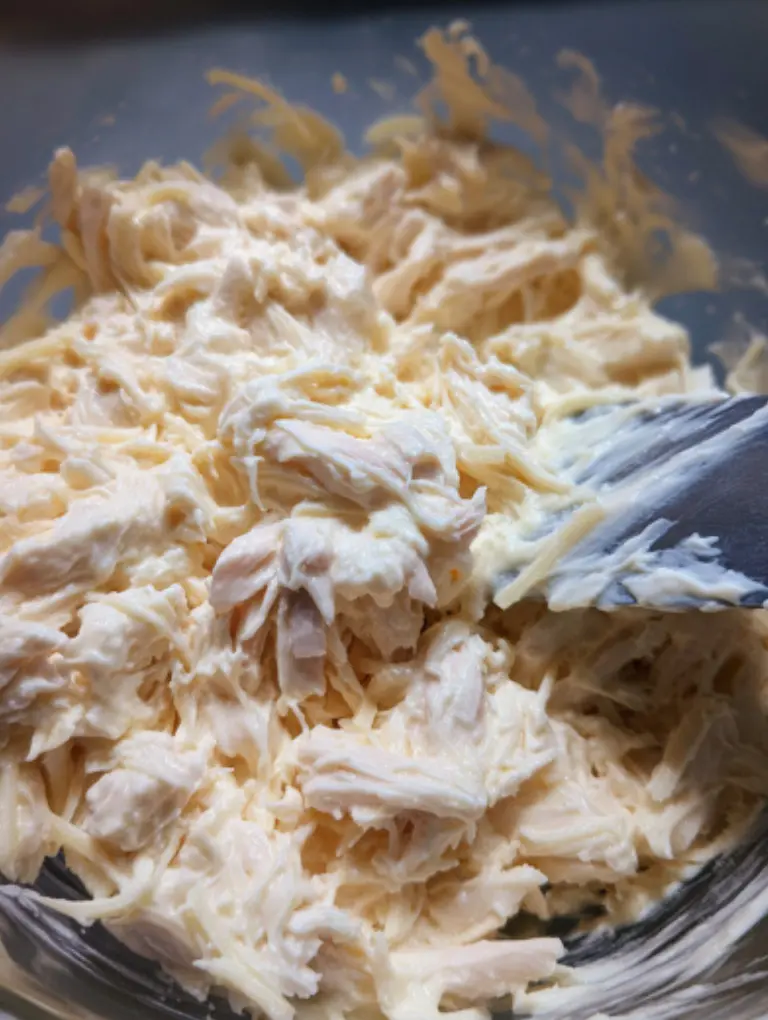Overhead view of a bowl mixing shredded chicken, garlic, mayonnaise, Greek yogurt, and melted cheese with a spatula, creamy and glossy texture on a kitchen counter.