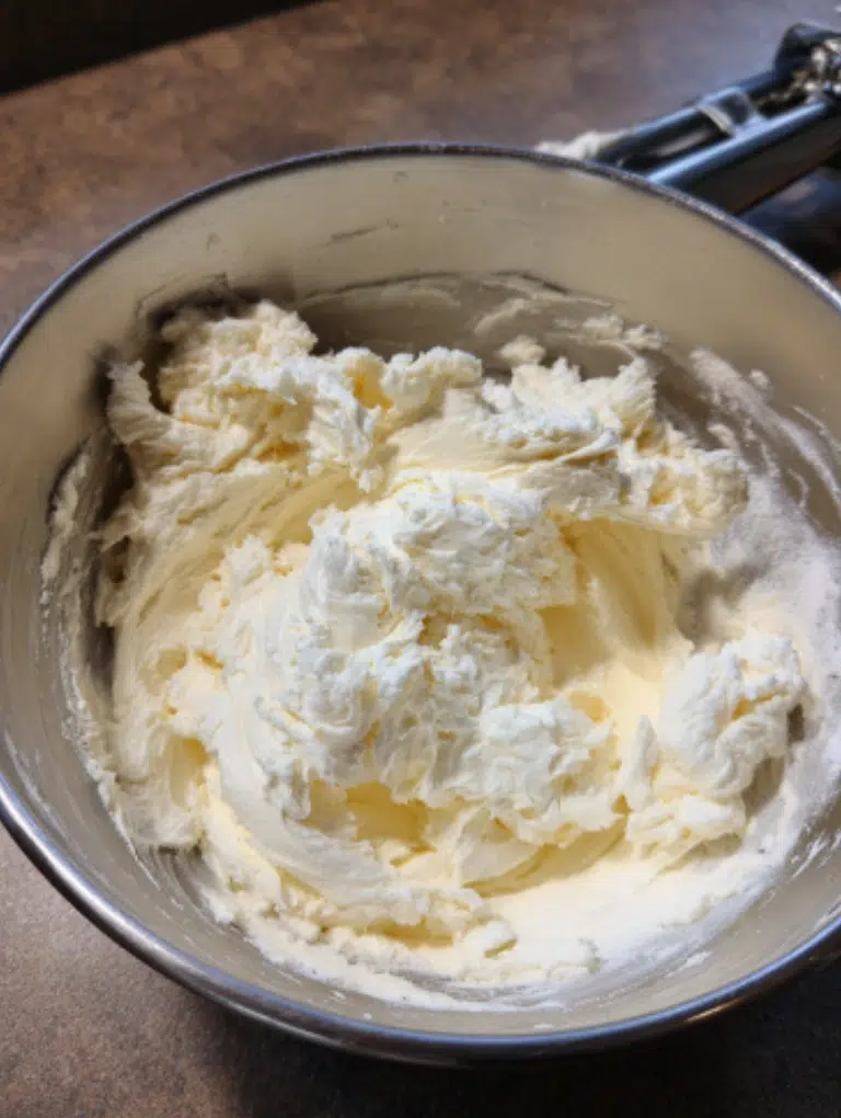 Cream cheese, sugar, and vanilla mixture being whipped in a bowl on a kitchen counter with a hand mixer.