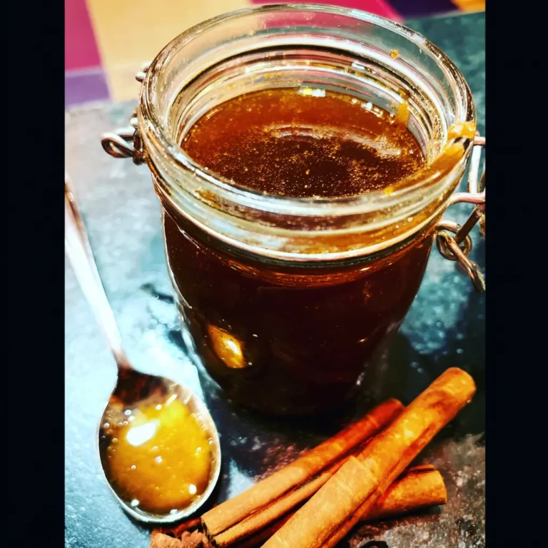 A rustic still life featuring a clear glass jar of homemade gingerbread syrup, a spoon with the viscous syrup, and aromatic cinnamon sticks on a dark, textured surface, hinting at a delicious gingerbread syrup recipe.